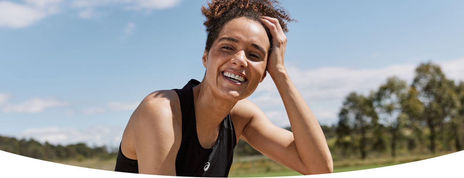 A female runner sits in the grass laughing and smiling toward the camera with her hand in her hair, wearing a black ASICS sleeveless top