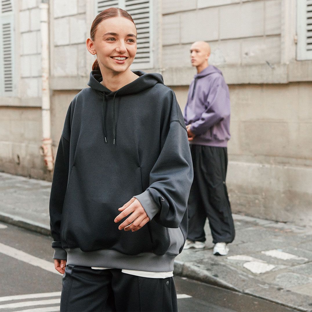 This tile image features a female model in the foreground wearing the new ASICS SportStyle apparel collection black hoodie. In the background, a male model wears the purple hoodie.