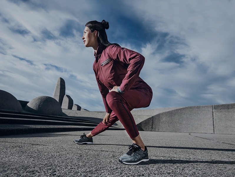 Woman in a red athletic jacket and pants and gray running shoes stretching outside.
